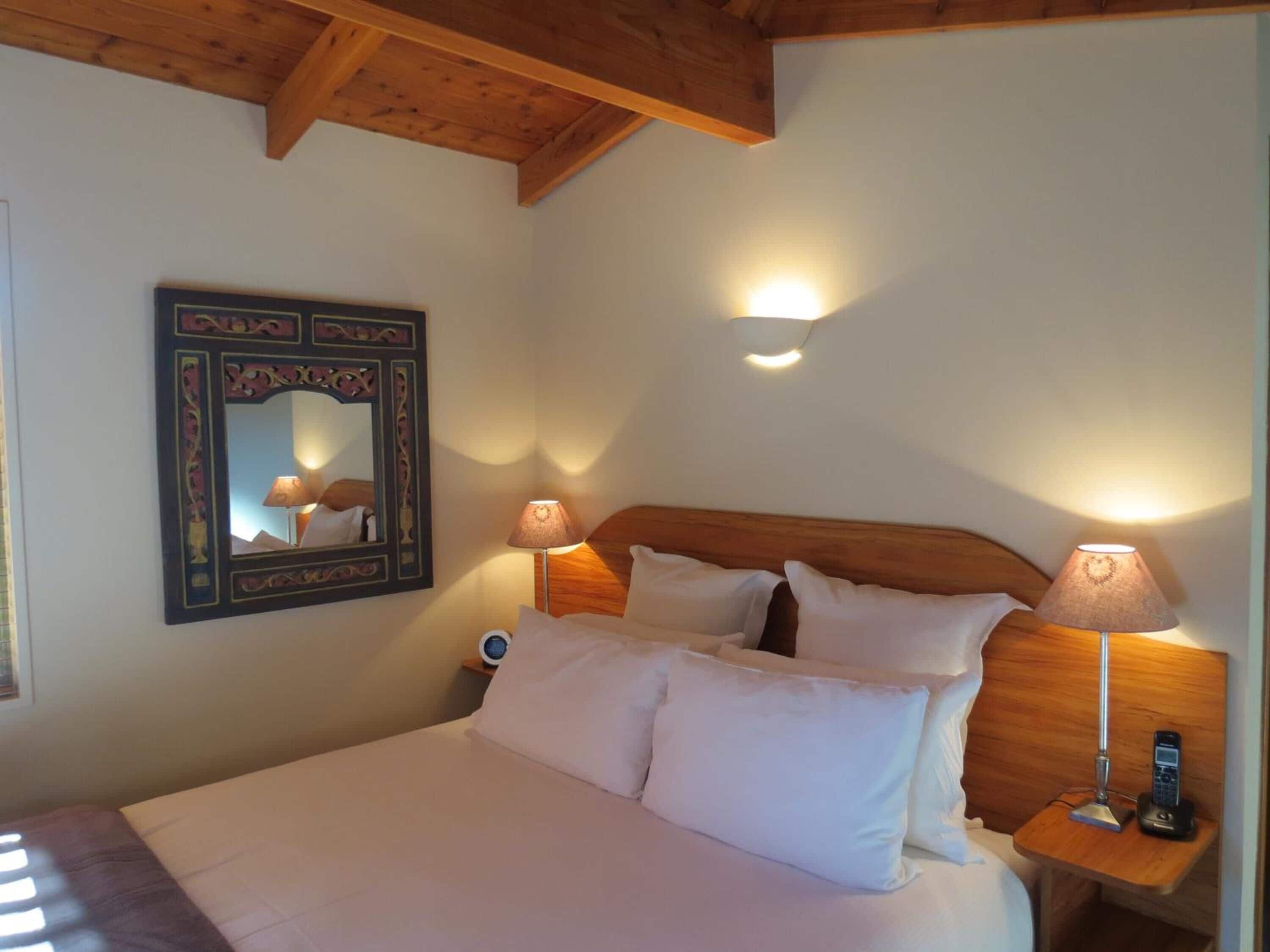 A bedroom with a wooden headboard, a bed with white linens, two bedside lamps, a wall-mounted mirror, and a clock on the nightstand.
