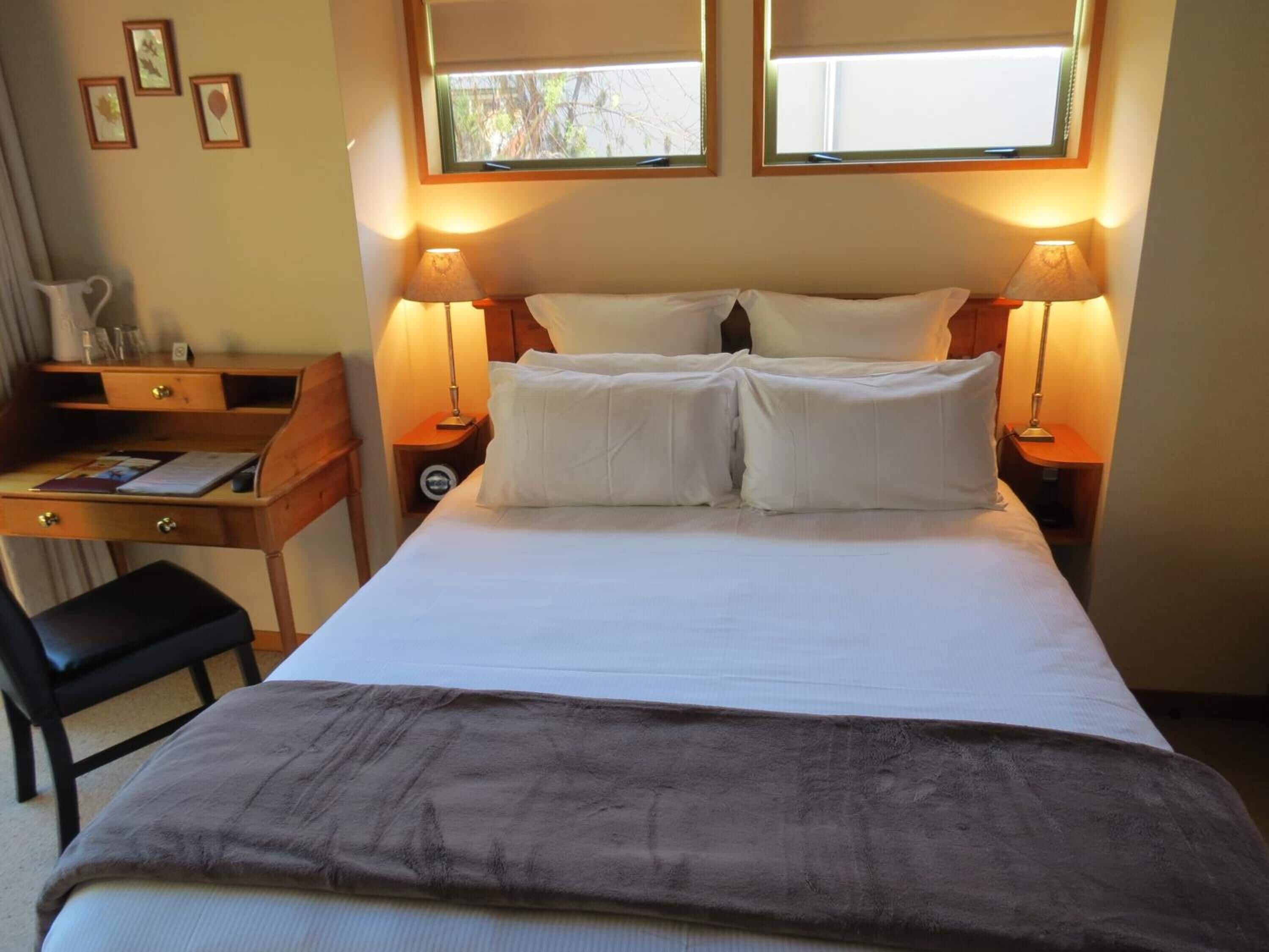 Una habitación de hotel con una cama, dos lámparas de noche, un escritorio con una silla y una ventana con persianas.