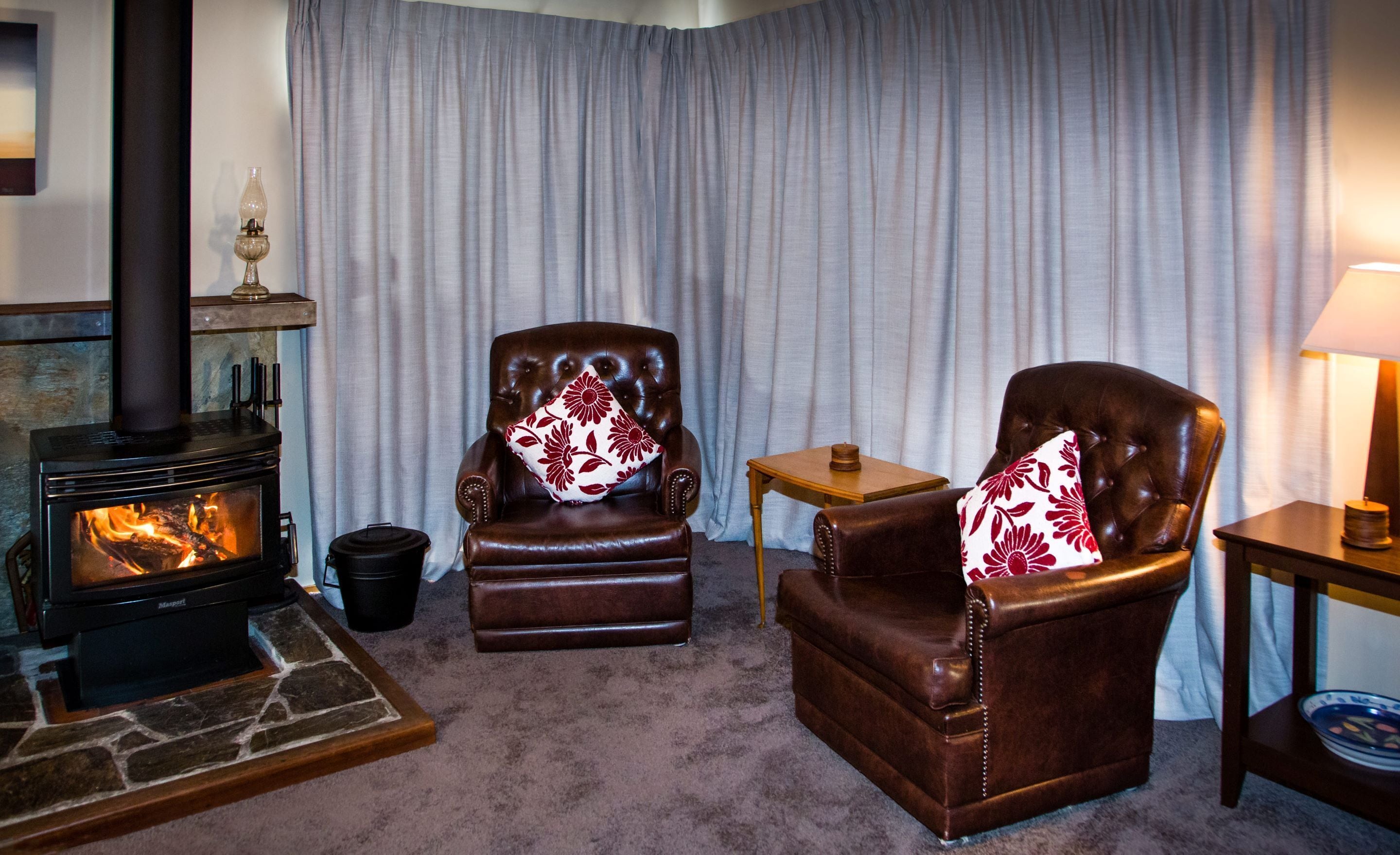Un salon chaleureux avec une cheminée, deux fauteuils en cuir ornés de coussins à motifs floraux, une petite table en bois et une lampe.