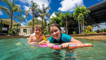 Two people in a swimming pool with palm trees and a building in the background.