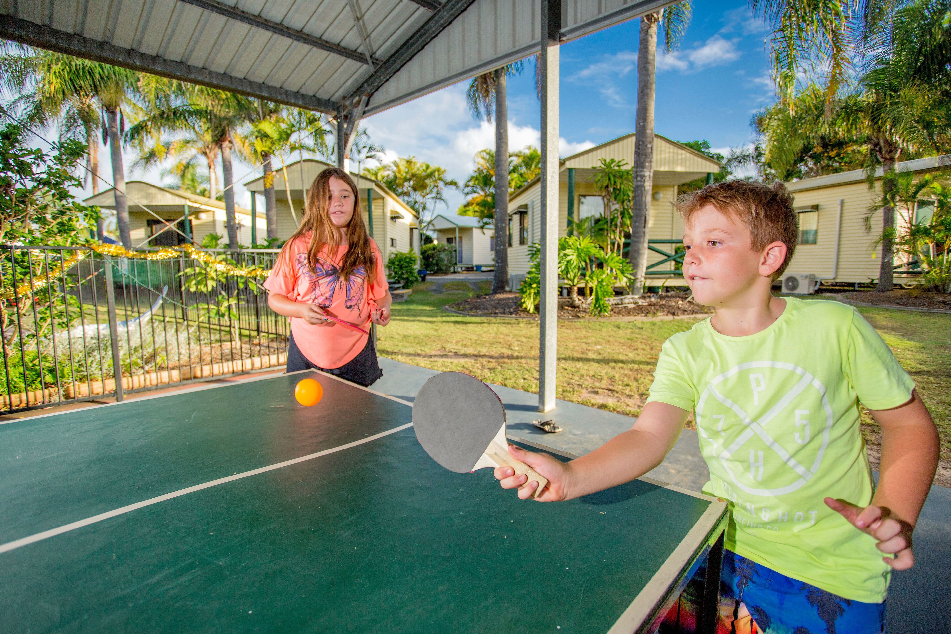 Una cancha de tenis con red, rodeada por una cerca metálica, palmeras y casas al fondo.