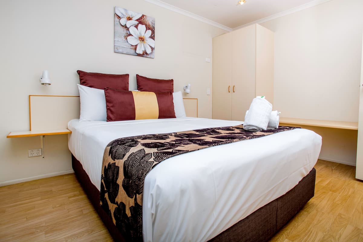 Una cama bien hecha con una colcha estampada, una obra de arte en la pared con flores blancas y una mesita de noche de madera con una lámpara.