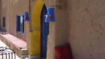 Decoración tradicional marroquí con una mesa lista para el desayuno, incluyendo pan, fruta y bebidas, frente a un edificio con un arco amarillo y el nombre “KASBAH” a la vista.