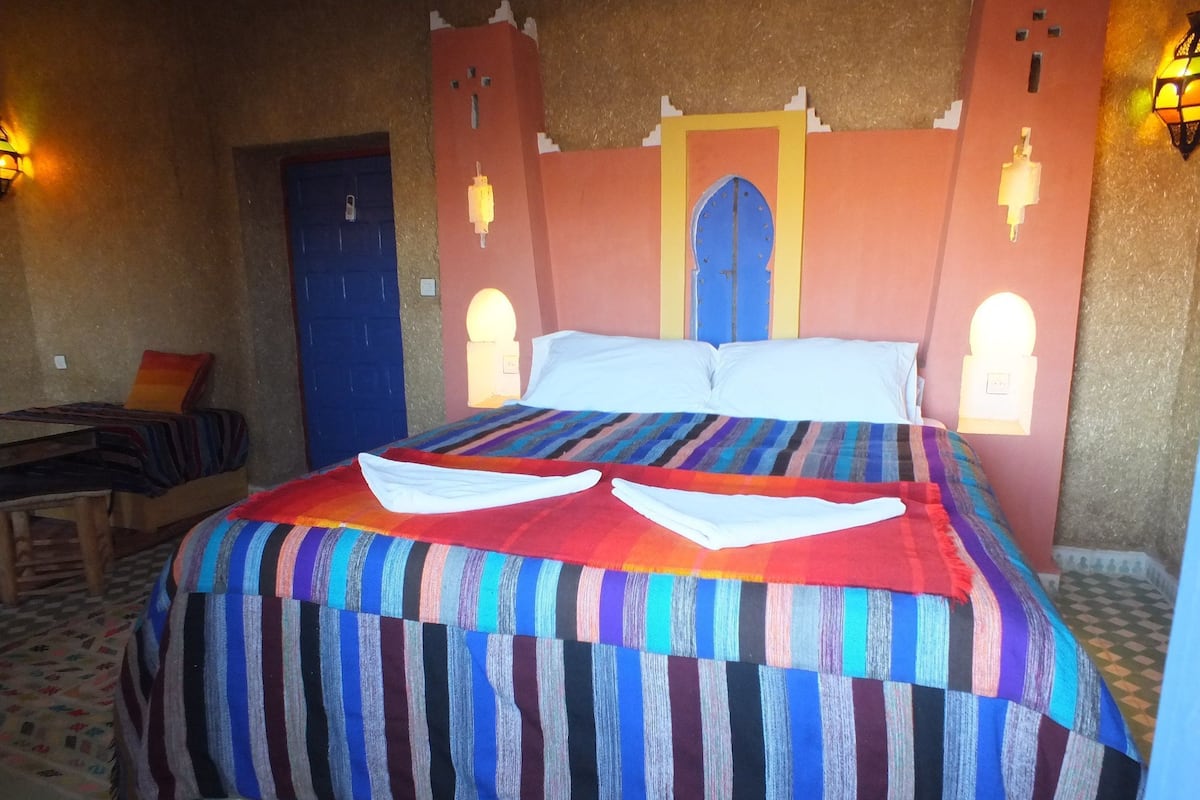 Una cama con una colcha a rayas de colores, una puerta azul y dos lámparas fijadas a la pared.