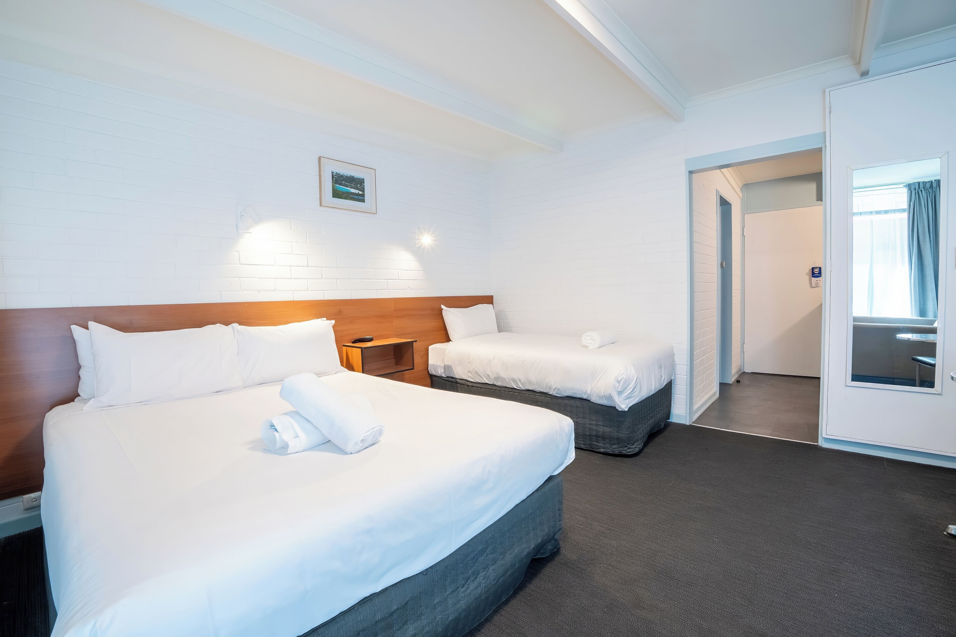 A hotel room with two beds, a wooden headboard, a nightstand, a framed picture on the wall, and a door leading to a bathroom.