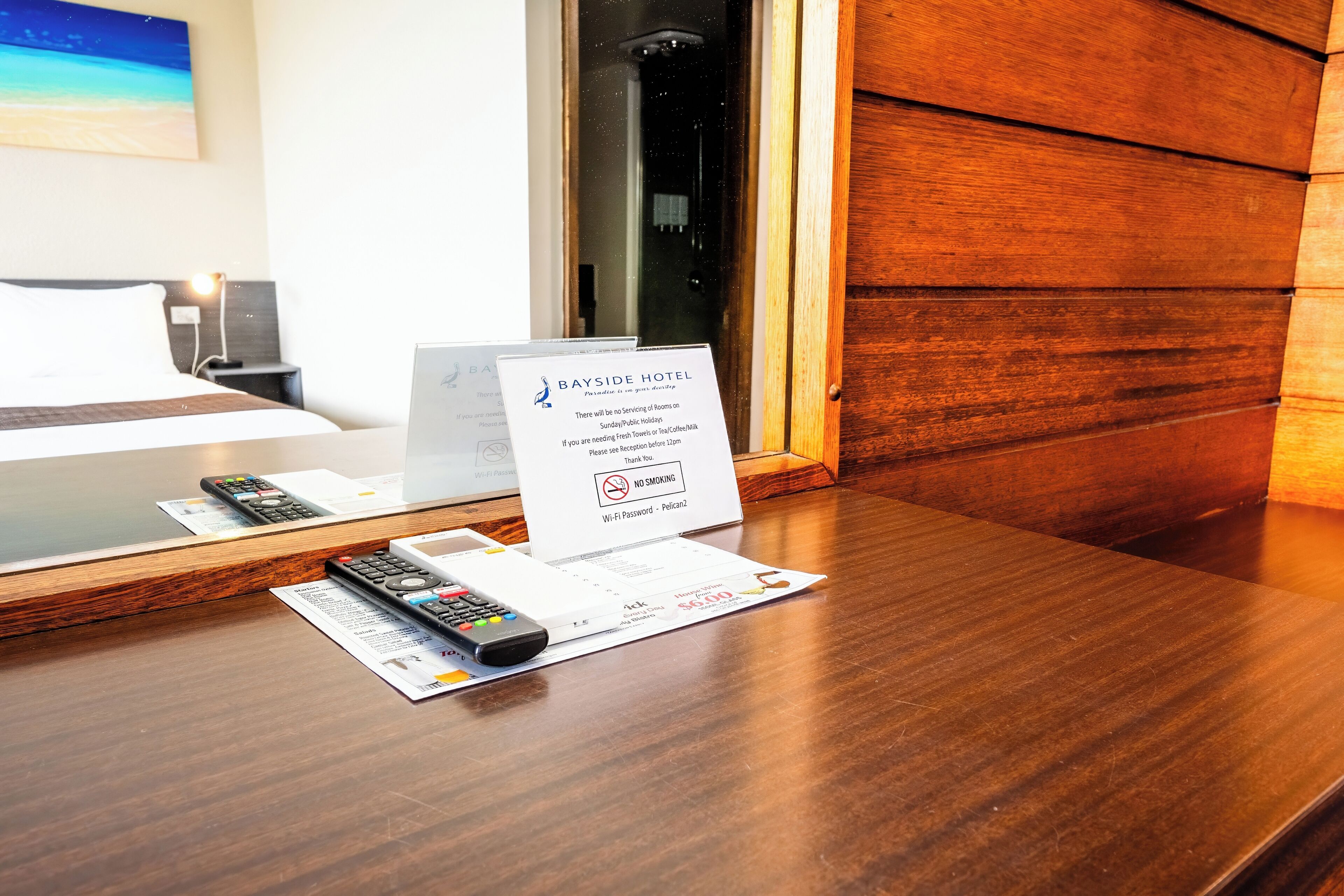 Een hotelkamer met een houten bureau, een bed, een bord met informatie over roomservice en een raam met uitzicht op het strand.