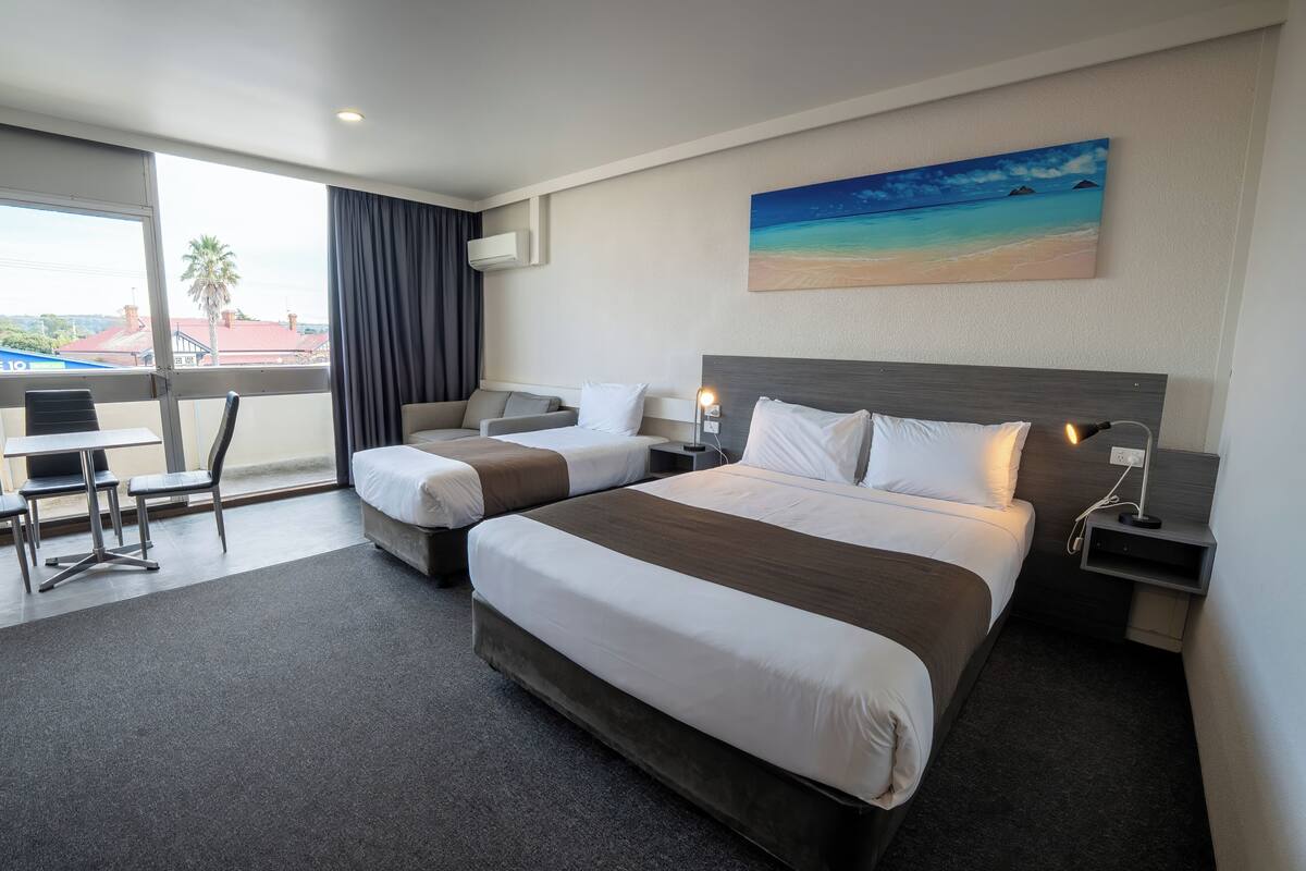 A hotel room with two beds, a desk, and a large window with a view of buildings and palm trees.