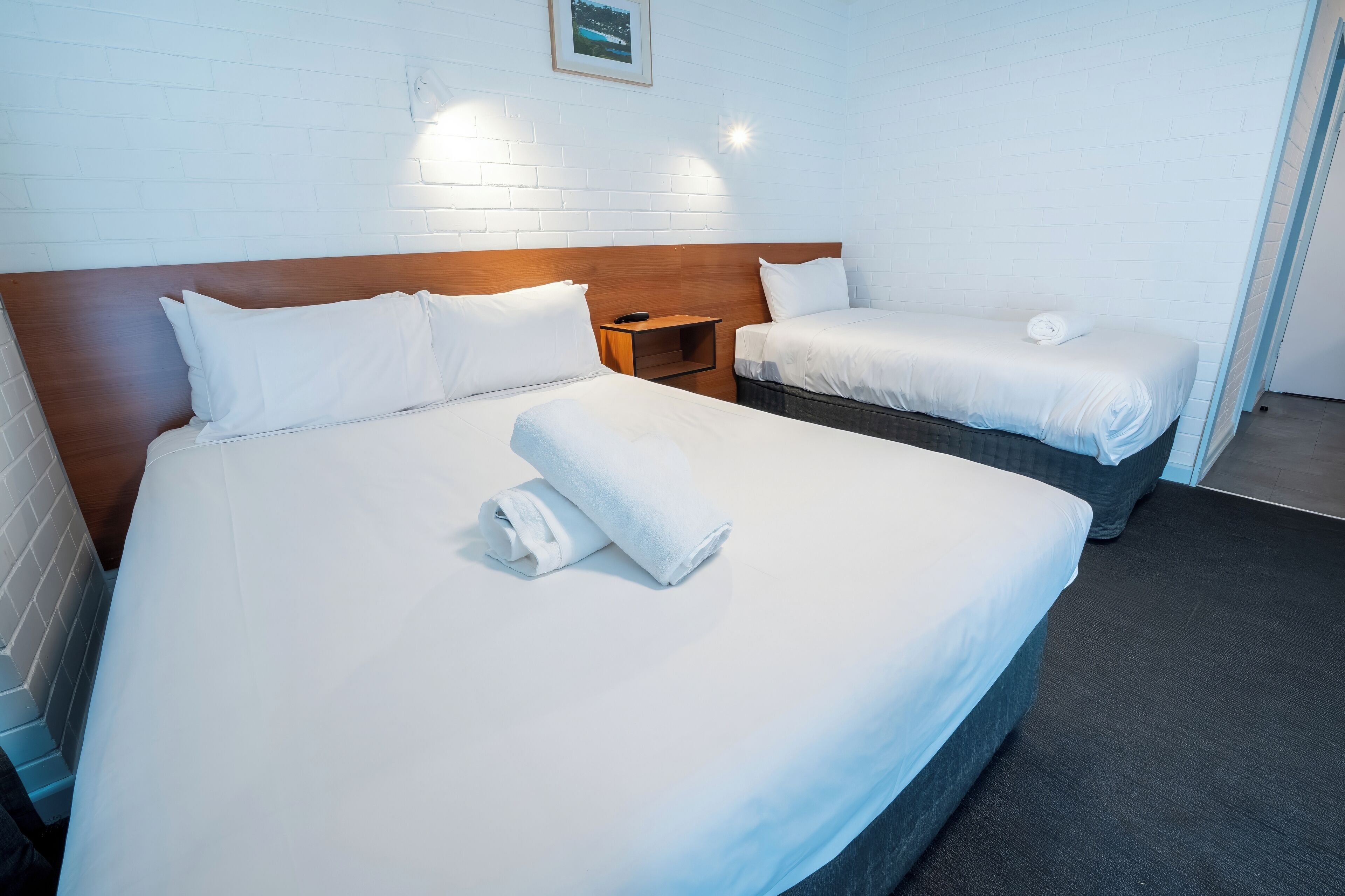 Habitación de hotel con dos camas, sábanas blancas, cabecero de madera, una mesita de noche y un cuadro en la pared.