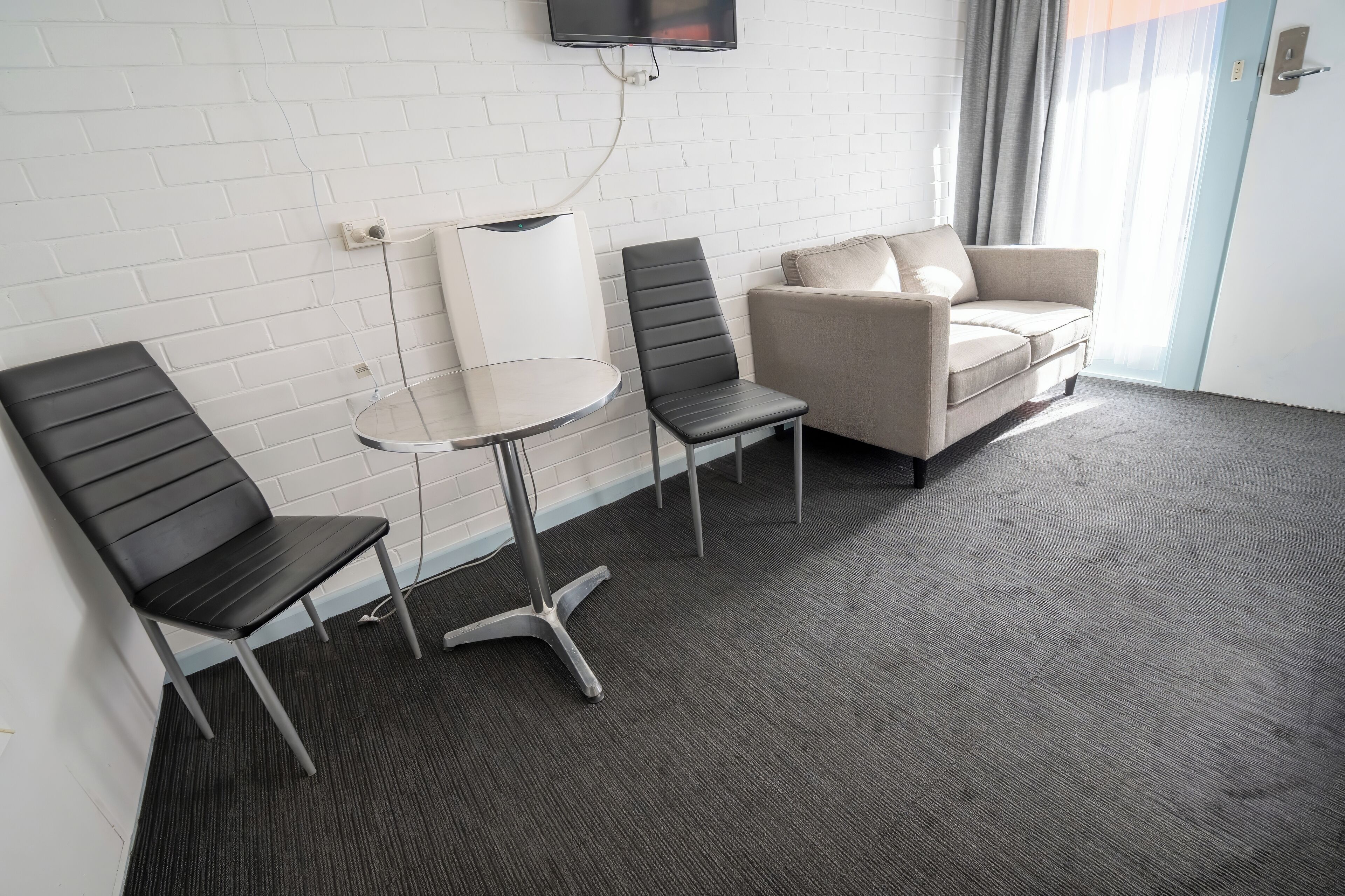 Hotelkamer met een witte baksteenmuur, een flatscreen televisie boven een witte radiator, een grijs vloerkleed, een beige bank en twee zwarte stoelen.