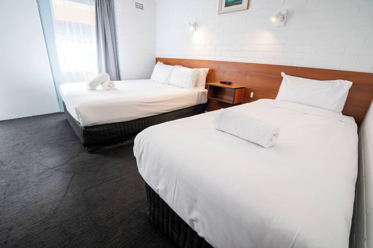 A hotel room with two beds, a wooden headboard, a nightstand, and a window with curtains.