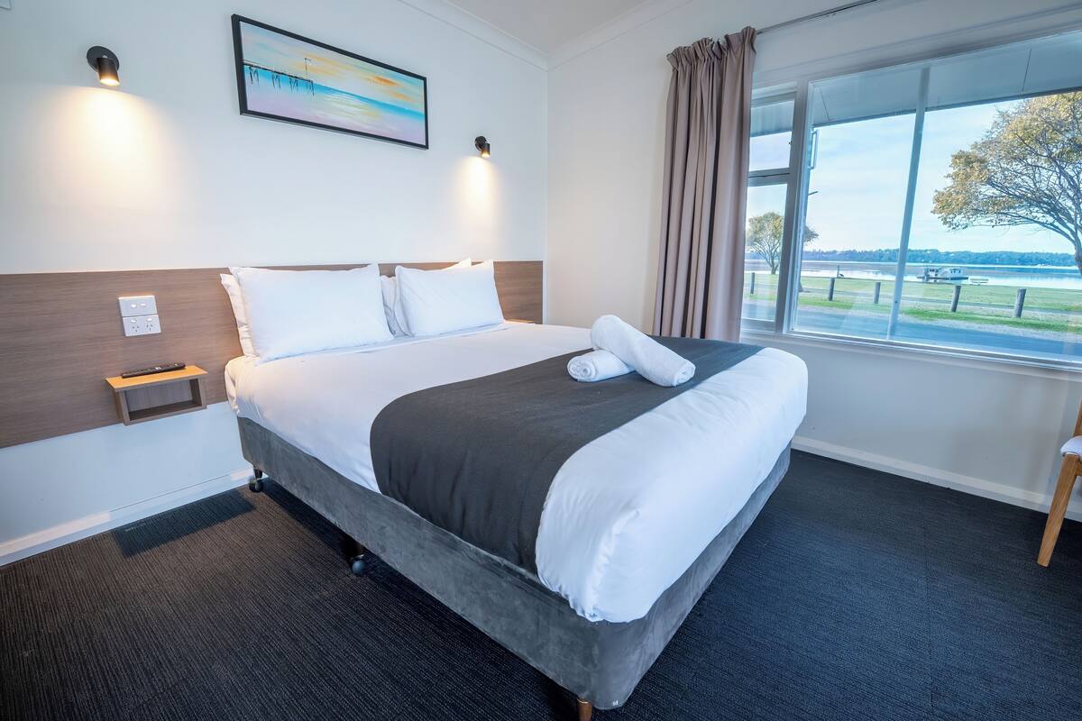 A hotel room with a bed, a nightstand, a remote control, a painting, and a window with a view of a park and lake.