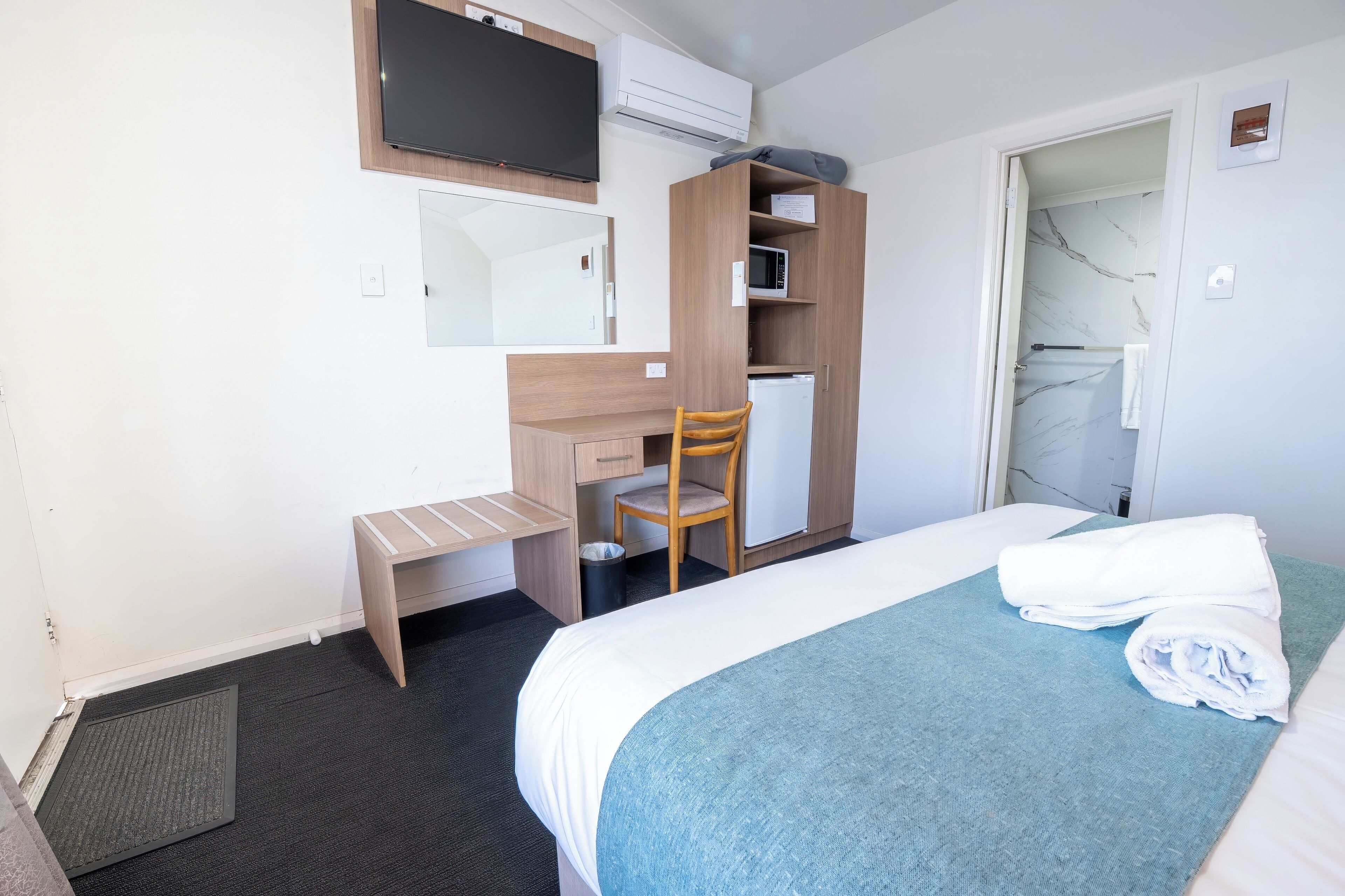 A hotel room with a bed, a desk, a chair, a wardrobe, and a bathroom visible through an open door.