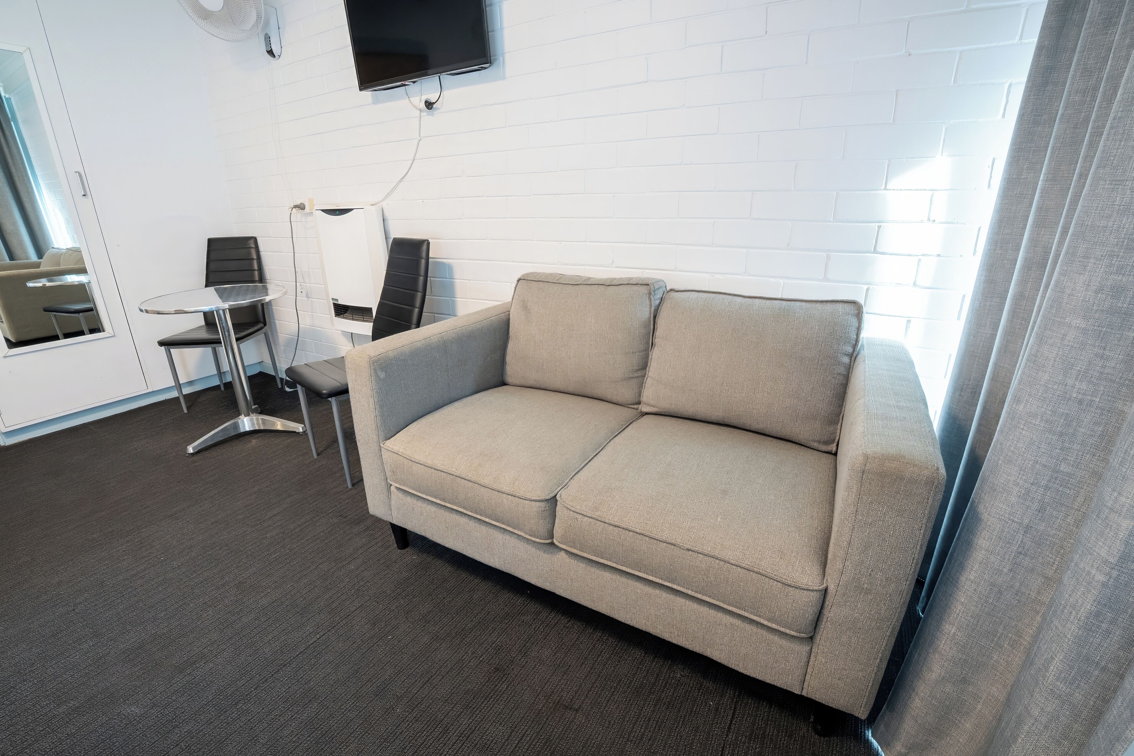 A modern living room with a beige sofa, a glass table, and two chairs. A flat-screen TV is mounted on the wall, and a window with curtains is visible.