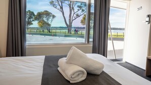 Una habitación de hotel con una cama, un ventanal con vista a un parque y un lago, y una puerta con el número 303.