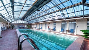 Una piscina interior con techo de cristal, suelo de baldosas y zona de estar con sillas y plantas en macetas.