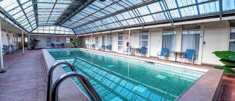 Una piscina interior con techo de vidrio, sillas de piscina y suelo de baldosas.