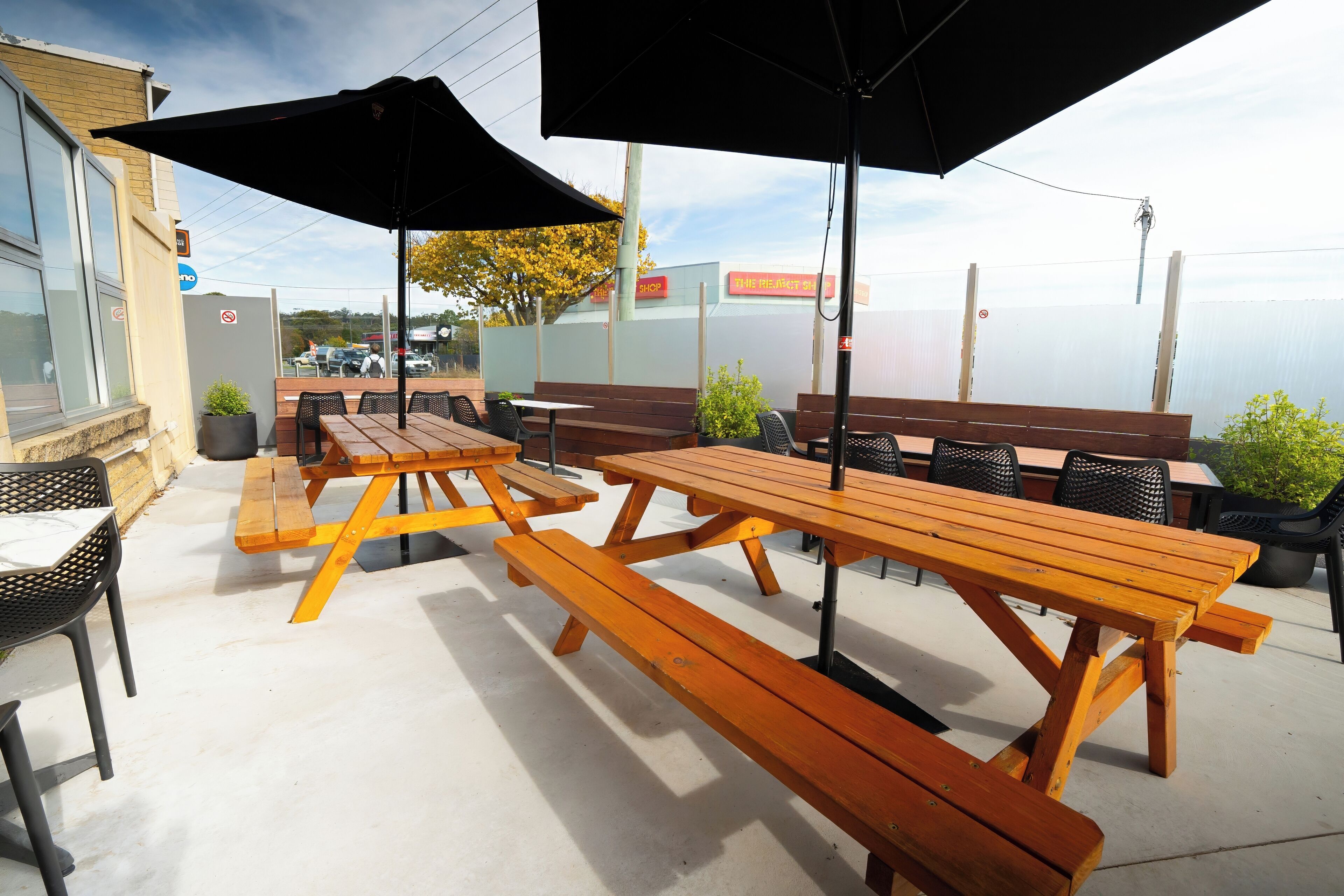Un patio al aire libre con mesas y bancos de picnic de madera, sombrillas negras y plantas en macetas.