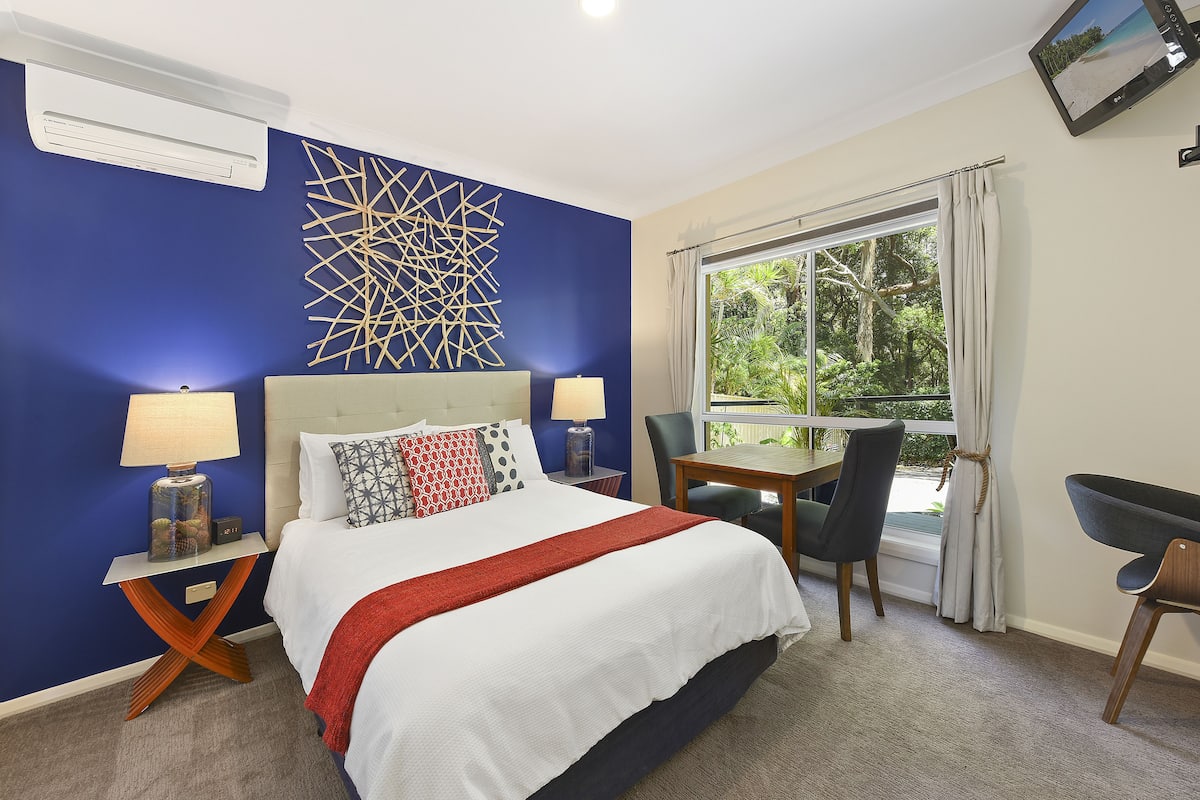 Une chambre d’hôtel équipée d’un lit, d’un bureau, d’une chaise, d’une fenêtre avec des rideaux et d’un téléviseur fixé au mur.