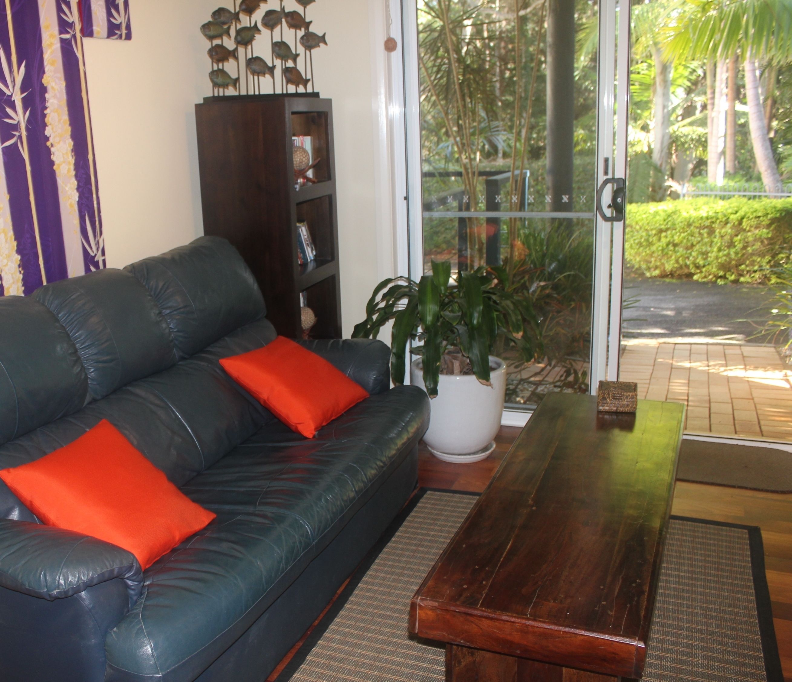 A living room with a black leather sofa, red cushions, a wooden coffee table, a white potted plant, and a bookshelf.