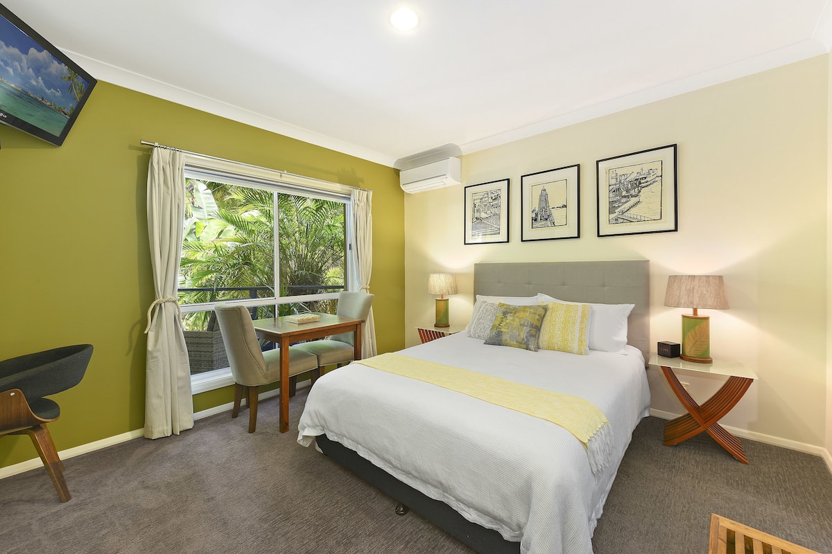 Une chambre d’hôtel avec un lit, un bureau et une chaise. Il y a une fenêtre avec des rideaux, une lampe et des tableaux encadrés au mur.