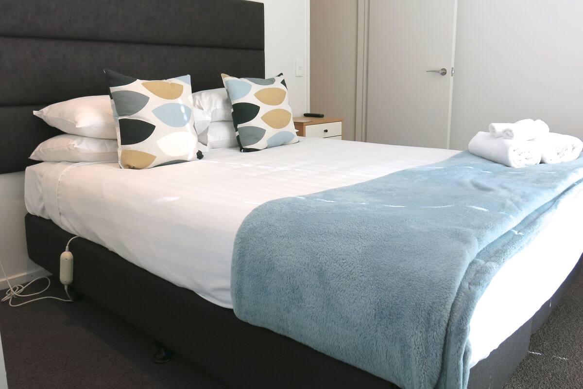 Un lit bien fait, avec une tête de lit sombre, une literie blanche et un plaid bleu.