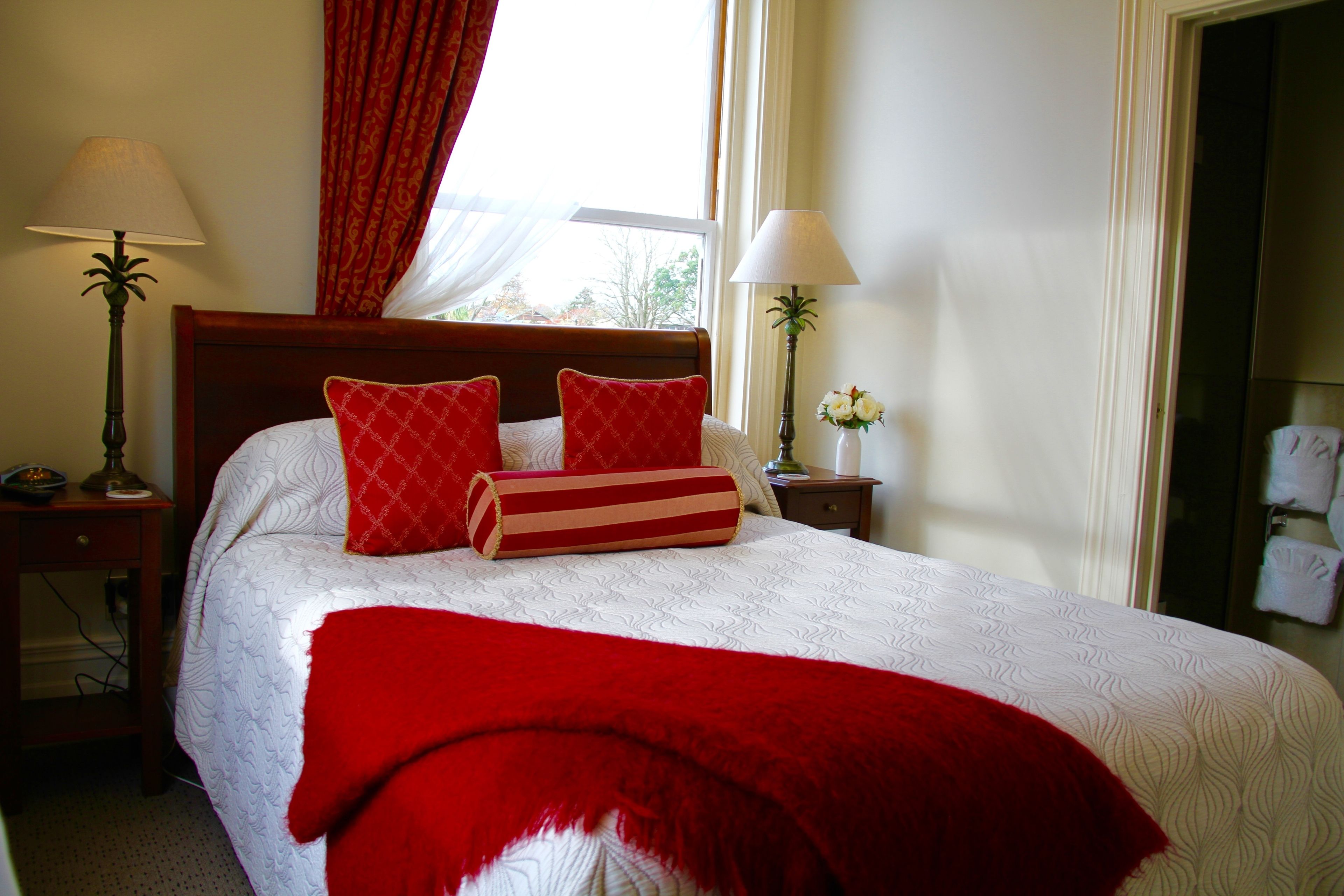 A hotel room with a large bed, red and white bedding, two bedside lamps, and a window with curtains.