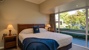 A hotel room with a large bed, two bedside tables, a lamp on each table, and a view of a garden through a large window. - Merivale Manor (Christchurch)