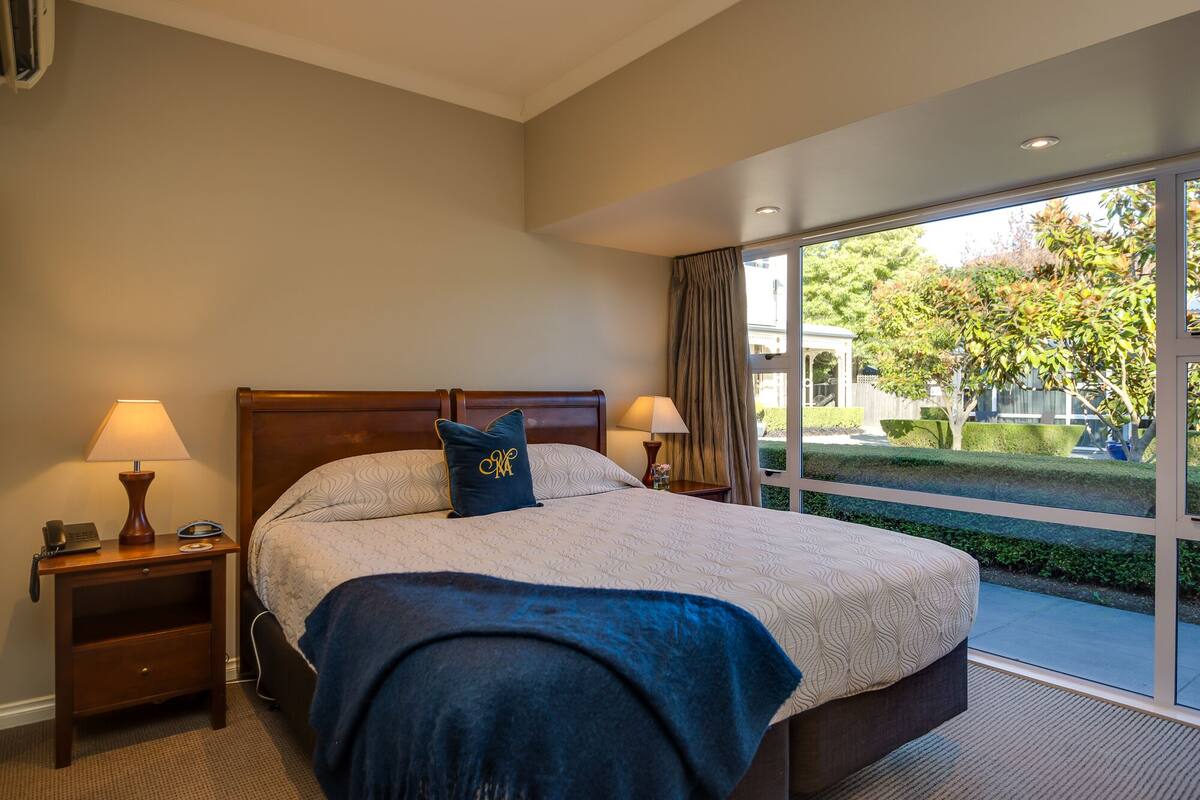 Une chambre d’hôtel avec un grand lit, deux tables de chevet, une lampe sur chaque table et une vue sur un jardin, visible à travers une grande fenêtre.
