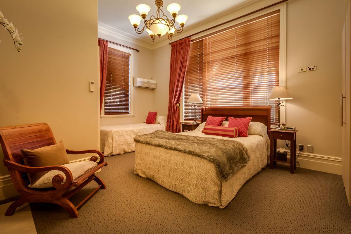 Une chambre d’hôtel avec deux lits, une chaise en bois, un lustre et de grandes fenêtres avec des rideaux.