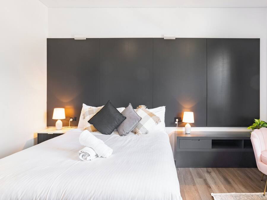 Et moderne hotelværelse med en stor seng, sengeborde med lamper, en mørk sengegavl og en lyserød stol.