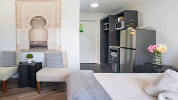 A modern hotel room with a bed, two armchairs, a small table with a plant, a decorative wall piece, a minibar, and a refrigerator.