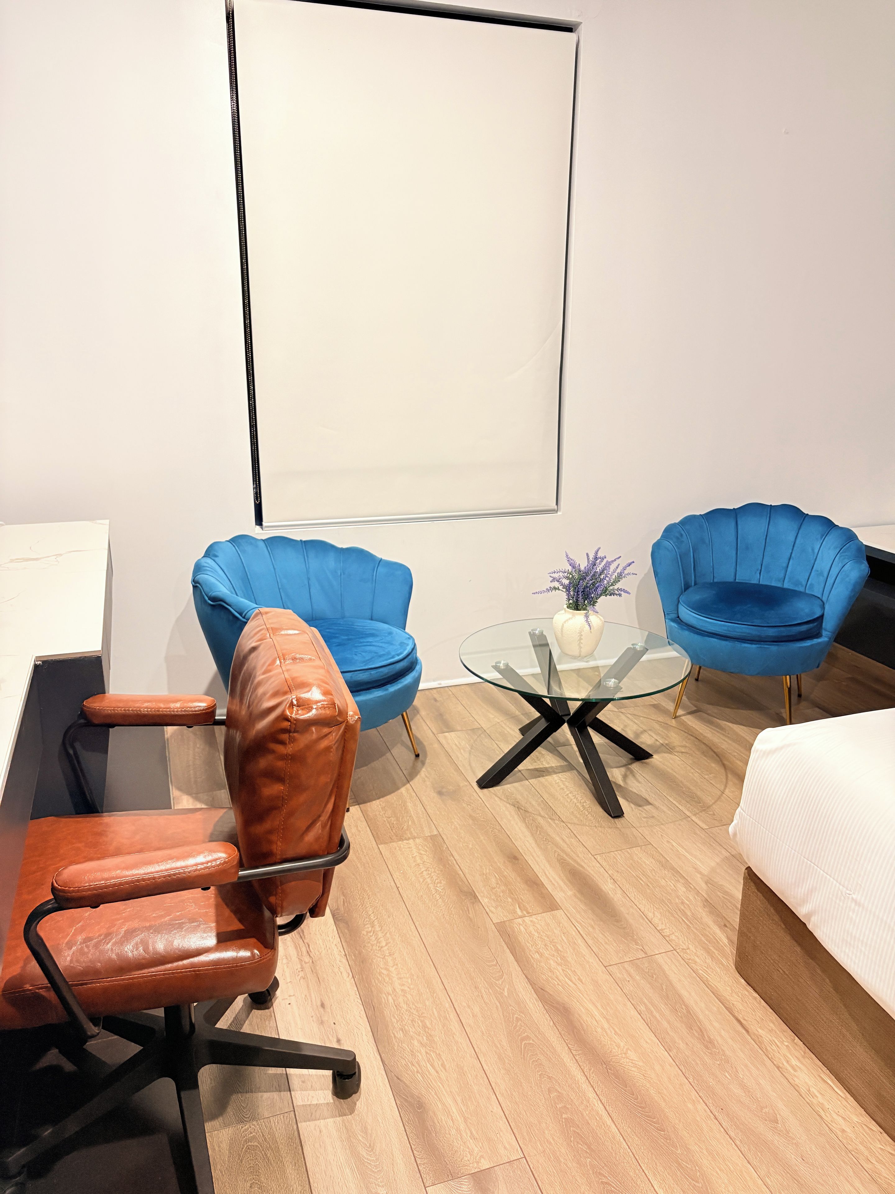 A modern room with a wooden floor, a glass table, two blue chairs, a brown leather chair, and a white bed.