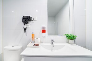 A bathroom with a white sink, a mirror, a hairdryer, a soap dispenser, a toilet, and a potted plant. - Bankstown Motel 10 (Greenacre)