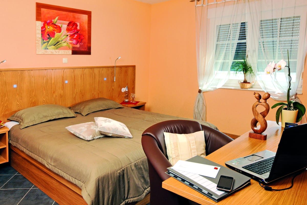 Une chambre avec un lit, un bureau avec un ordinateur portable, une chaise, une table de chevet, une plante en pot et une fenêtre avec des rideaux légers.