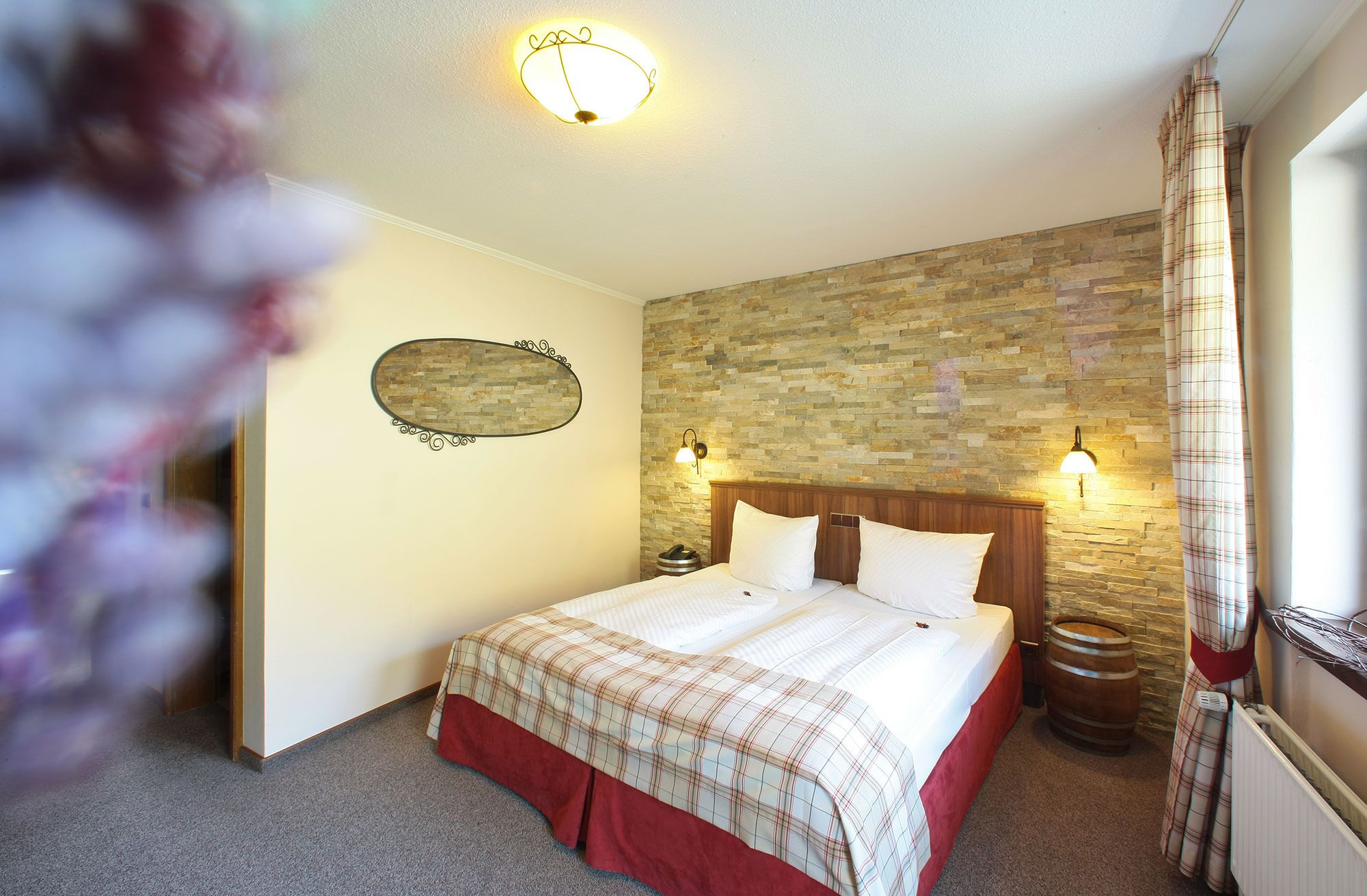 A bedroom with a stone wall, a wooden headboard, a bed with a plaid bedspread, two bedside lamps, a telephone, and a wooden barrel.