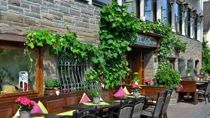 Un café en plein air avec des chaises et des tables en osier, un mur de pierre et une enseigne indiquant « EL Lellmann ».