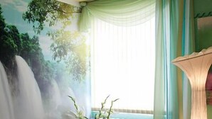 A room with a bed, a massage table, a wall mural, a window with curtains, and a floor lamp. - Fröbelhof (Bad Liebenstein)