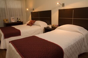 Habitación de hotel con dos camas, cada una con una manta roja estampada, una mesita de noche con un teléfono y una cesta, y una lámpara de pared.