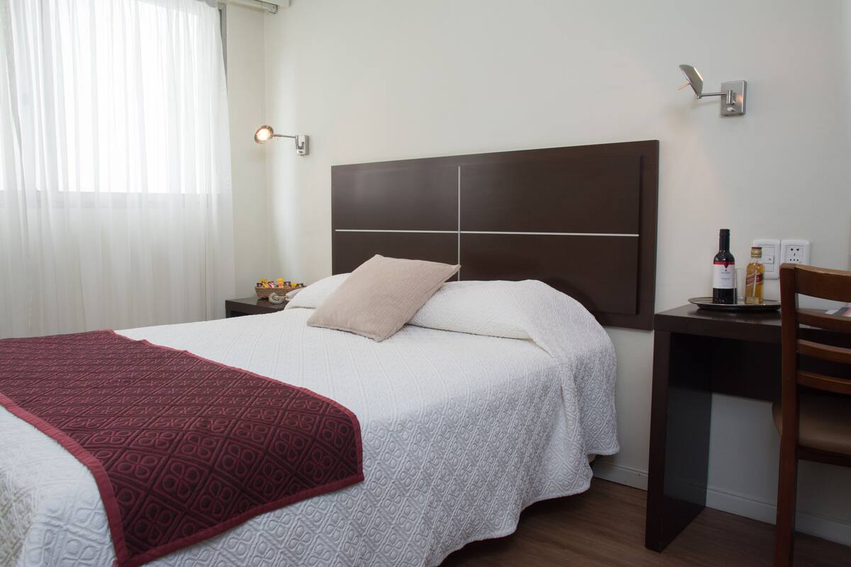 A hotel room with a large bed, a bedside table with a bottle and glasses, a desk with a chair, and a window with sheer curtains.