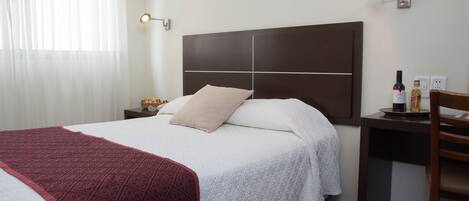 Ett hotellrum med en stor säng, ett nattduksbord med en flaska och glas, ett skrivbord med en stol och ett fönster med tunna, genomskinliga gardiner.