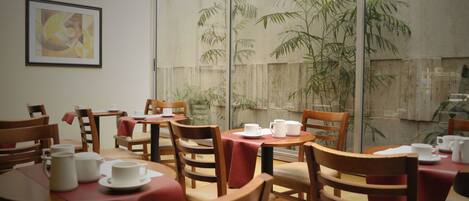 Un comedor con mesas y sillas de madera, un cuadro abstracto enmarcado en la pared y un amplio ventanal con vistas a plantas de bambú.