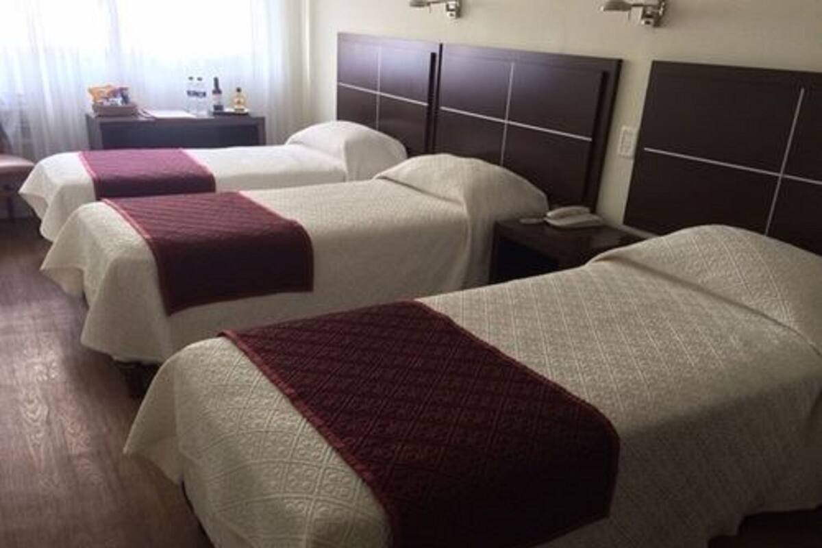 A hotel room with two beds, each with a white bedspread and a red blanket. There is a nightstand with a lamp and a bottle on it.