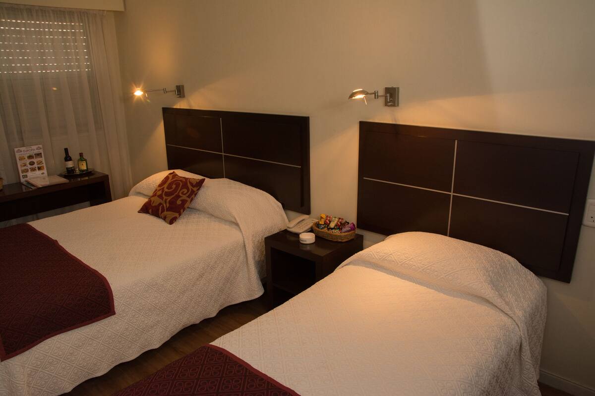 A hotel room with two beds, each with a headboard, bedside tables, and a nightstand with a lamp.