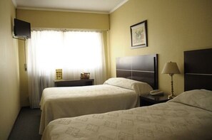 Habitación de hotel con dos camas, un televisor montado en la pared, una mesita de noche con lámpara y una ventana con cortinas.