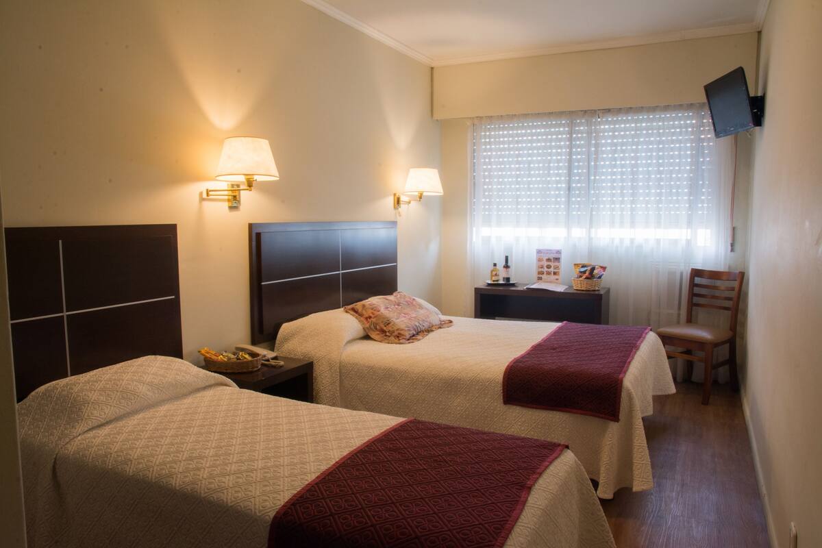 A hotel room with two beds, a TV mounted on the wall, a window with blinds, and a chair.