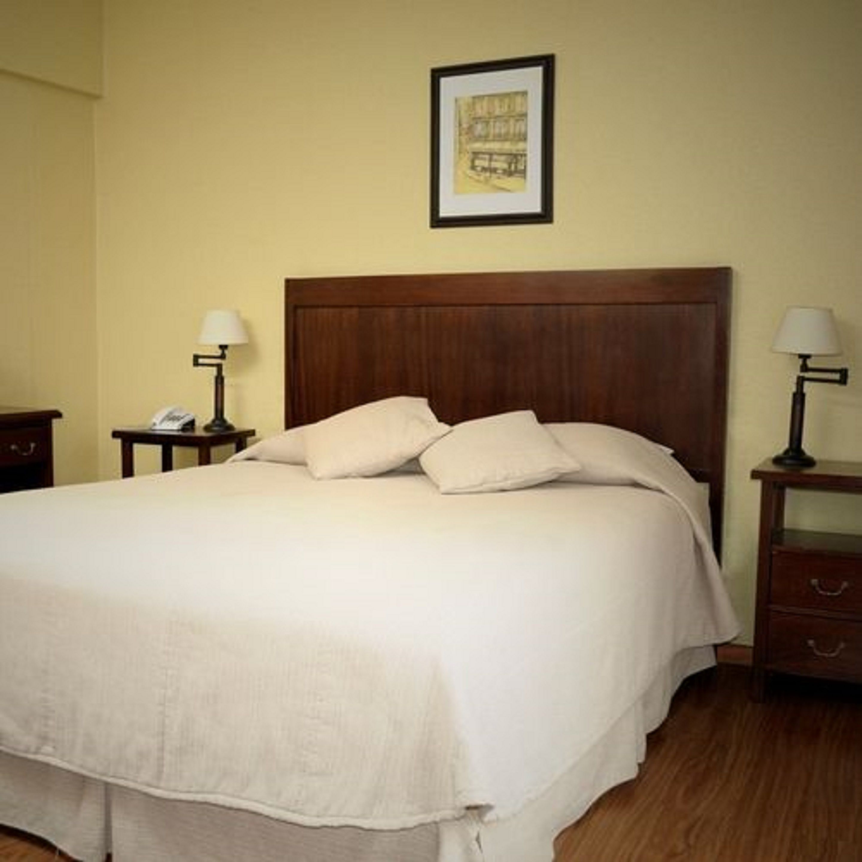 Una cama bien hecha con cabecera de madera, dos mesitas de noche con lámparas y un cuadro en la pared.