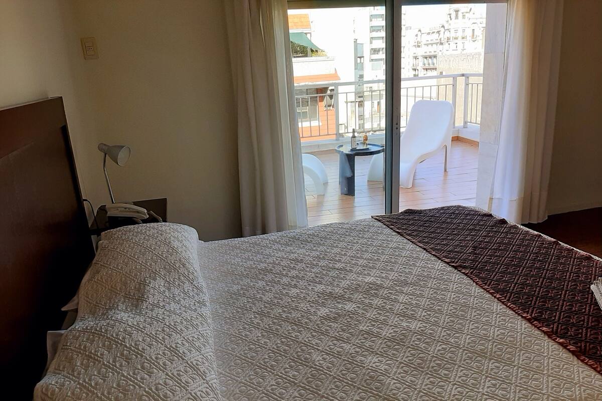 A hotel room with a large bed, a desk with a lamp, and a view of the balcony and outside buildings.