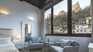A hotel room with a bed, a grey armchair, a sofa, a small table with a bottle and glass, and a large window offering a view of a historic building and trees. - Shine Albayzín Hotel (Granada)
