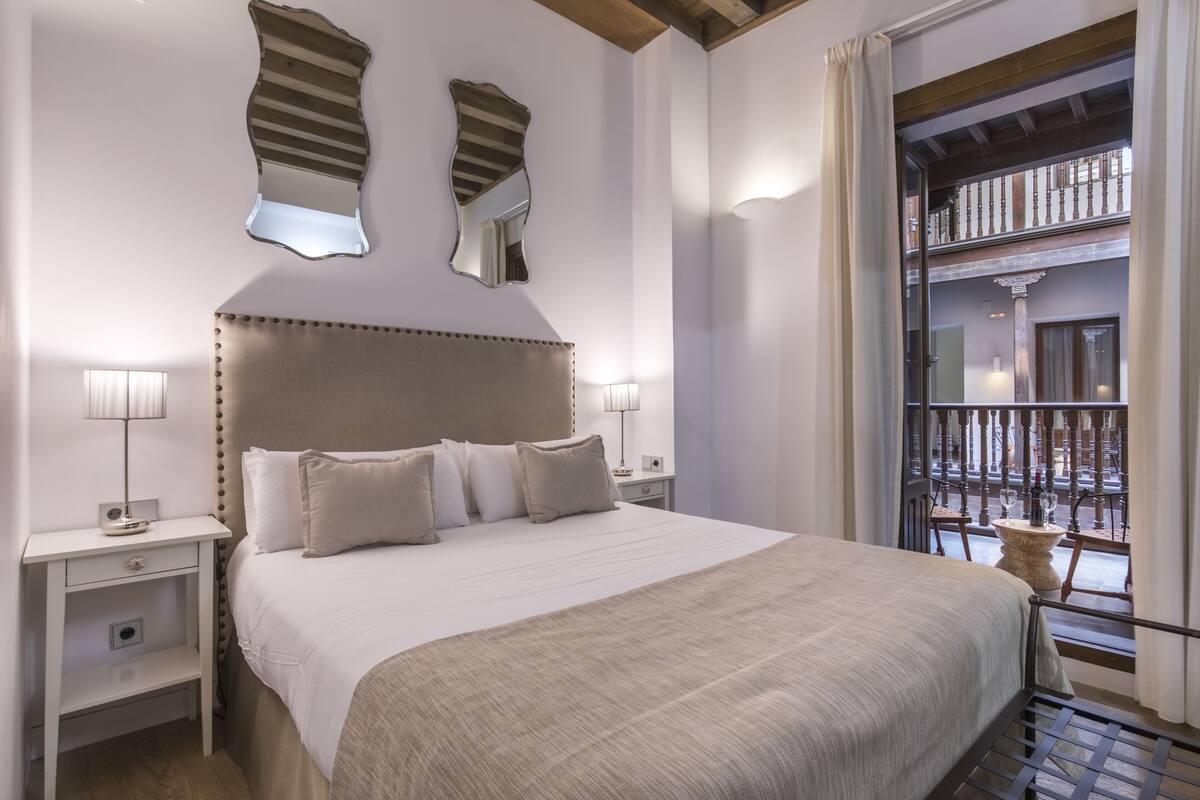 Habitación de hotel con una cama grande, dos mesitas de noche con lámparas, un espejo decorativo y vistas al balcón exterior.