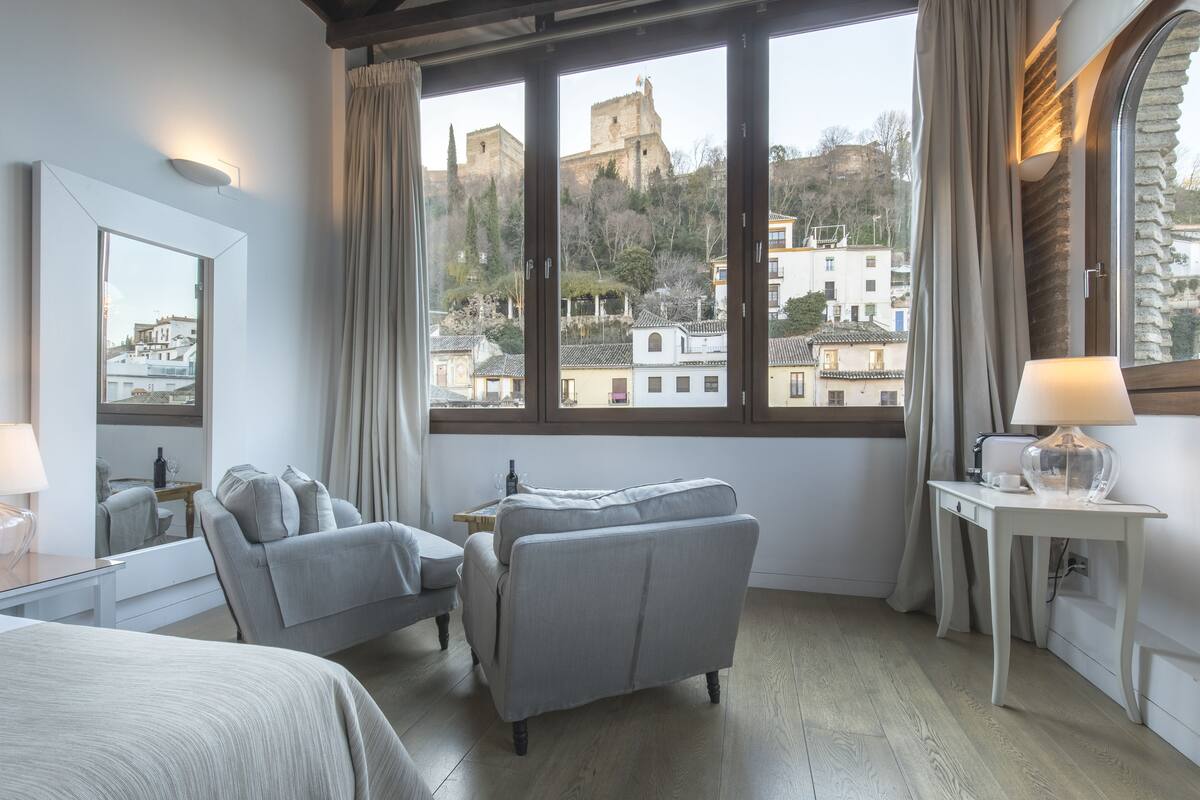 Habitación de hotel con una ventana grande, cama, sofá, una mesita de noche con lámpara y espejo.